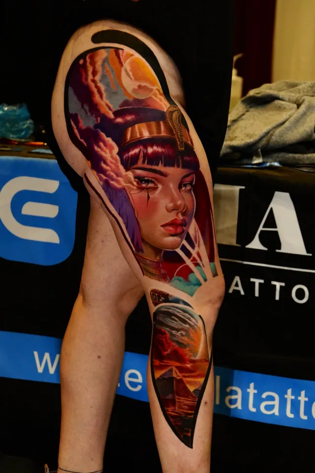 Color realism leg tattoo of an Egyptian queen with a golden cobra crown and violet clouds, with pyramids at sunset on the calf.