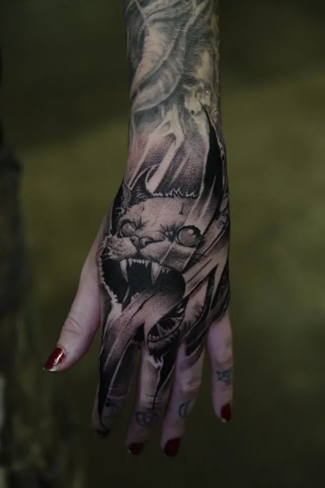 Black and gray hand tattoo of a hissing cat with fangs, surrounded by smoke and black brushstrokes.