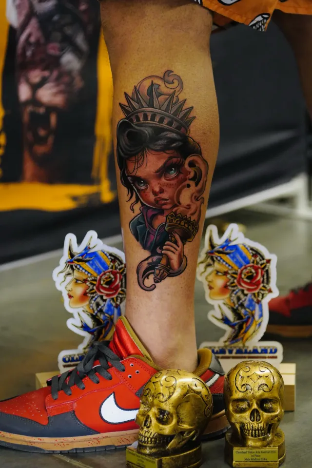 Neotraditional calf tattoo of a girl as the Statue of Liberty holding a torch, with two golden skull trophies on the floor.